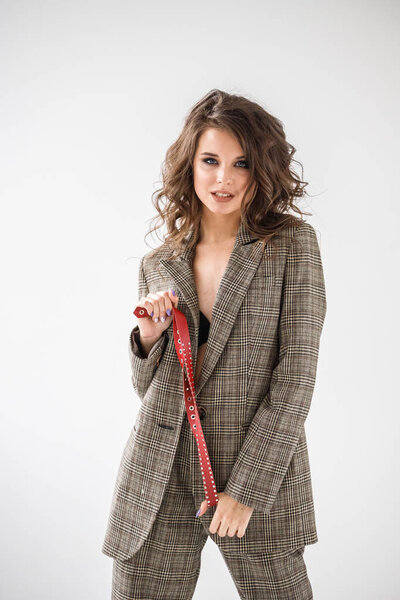 Fashion studio portrait of beautiful curly woman wearing checkered casual suit and black bra, standing on grey wall background with red belt in hands