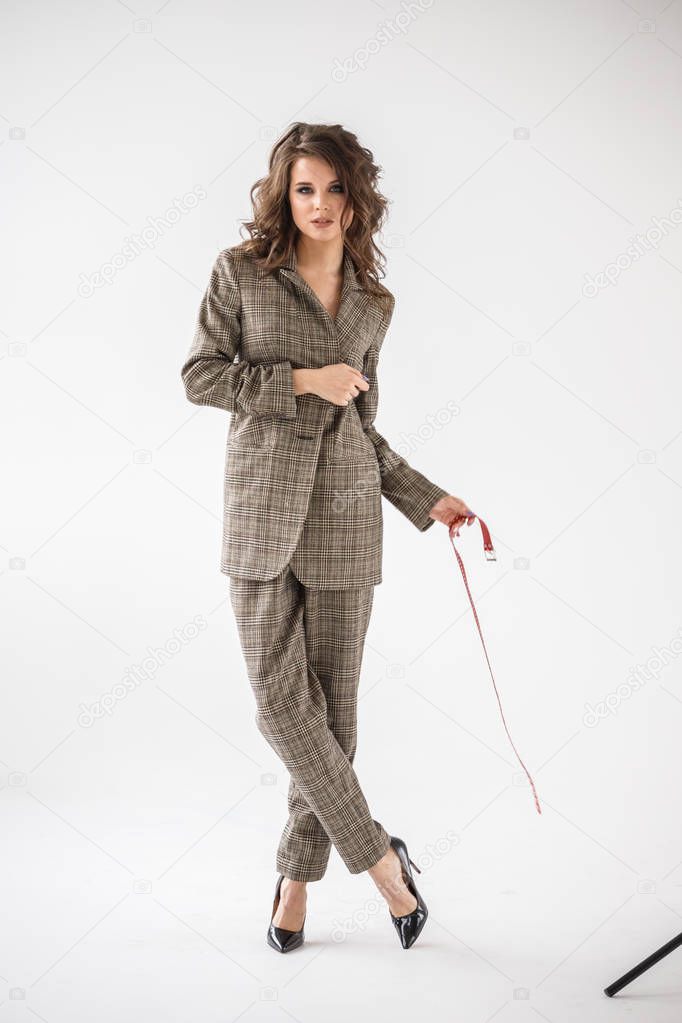 Fashion studio portrait of beautiful curly woman wearing checkered casual suit and black bra, standing on grey wall background with red belt in hands