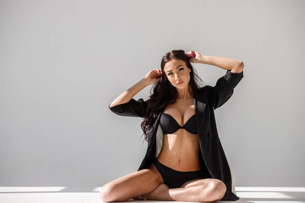Studio portrait of young brunette girl wearing sexy black lingerie and black coat sitting on floor, white wall background