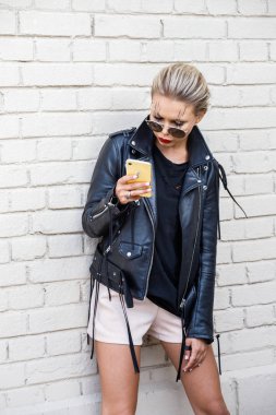 Siyah ceketli, gözlüklü, beyaz tuğla duvarda poz veren güzel sarışın kız. 