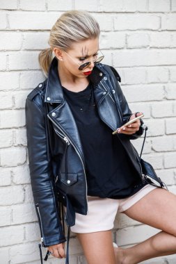 Siyah ceketli, gözlüklü, beyaz tuğla duvarda poz veren güzel sarışın kız. 