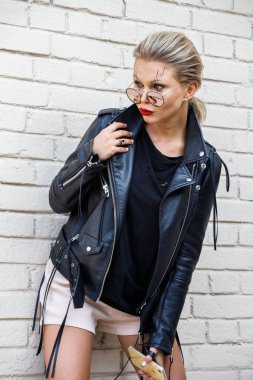 Siyah ceketli, gözlüklü, beyaz tuğla duvarda poz veren güzel sarışın kız. 