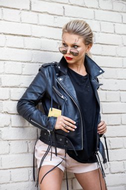 Siyah ceketli, gözlüklü, beyaz tuğla duvarda poz veren güzel sarışın kız. 