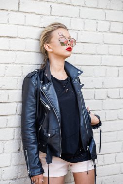 Siyah ceketli, gözlüklü, beyaz tuğla duvarda poz veren güzel sarışın kız. 