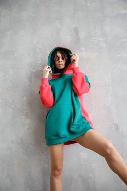 Güzel kız kapüşonlu seksi esmer duvarın yanında poz veriyor.