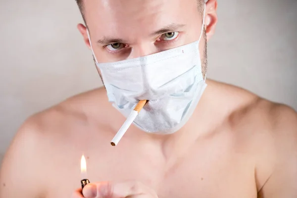 Mężczyzna bez ubrań w masce medycznej patrzy smutno w kamerę, trzymając papierosa w zębach. — Zdjęcie stockowe