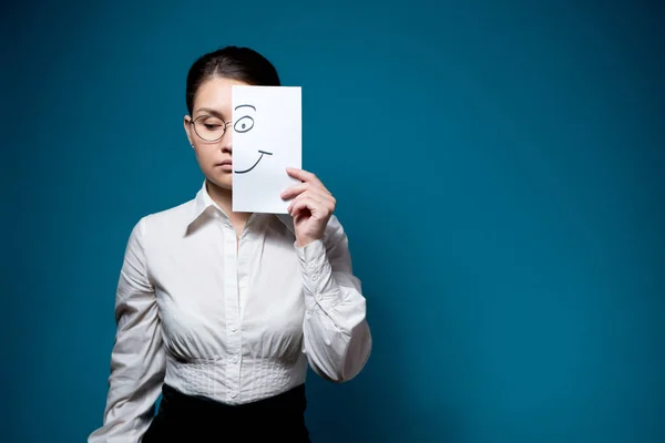 Unemotional woman with glasses and a white shirt hides her eyes and covers half her face with a piece o paper with a painted smile — Stock Photo, Image