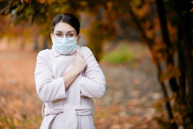 Tıbbi maskeli bir kız, sonbahar günü parktaki bir ceketin içinde donar.