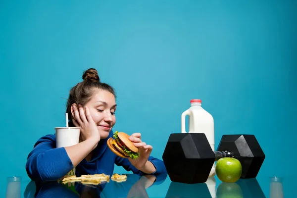 Het meisje met plezier kijkt naar de hamburger in haar hand terwijl voor haar ligt een sportuitrusting en gezond eten — Stockfoto