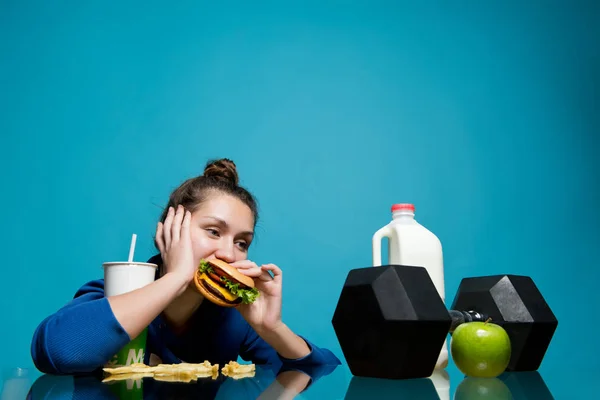 Het meisje kauwt lui op een hamburger en kijkt naar de halter, een fles melk en een appel — Stockfoto