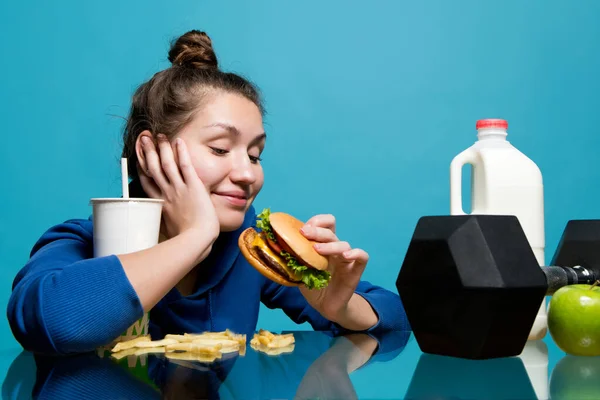 Дівчина дивиться на бургер з посмішкою, і перед нею лежить гантель — стокове фото