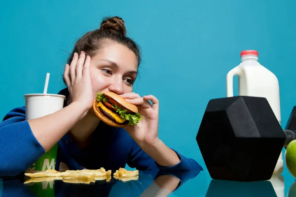 Jonge vrouw kauwt een hamburger lui en kijkt naar een halter — Stockfoto