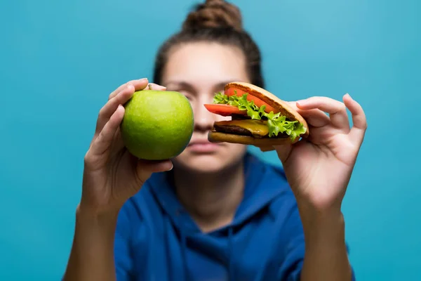 Дівчина тримає в руках бургер і яблуко поруч один з одним, вона сама знаходиться в зоні дефокусу — стокове фото