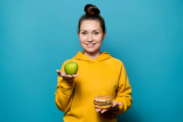 Meisje houdt een appel en een hamburger op de handpalmen met een glimlach — Stockfoto
