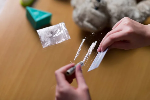 Γυναικεία χέρια ετοιμάζουν ίχνη κοκαΐνης, και στο παρασκήνιο είναι ορατά παιδικά παιχνίδια — Φωτογραφία Αρχείου