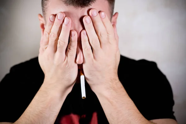 Man met een sigaret in zijn mond verlegen bedekt zijn gezicht met zijn handpalmen — Stockfoto