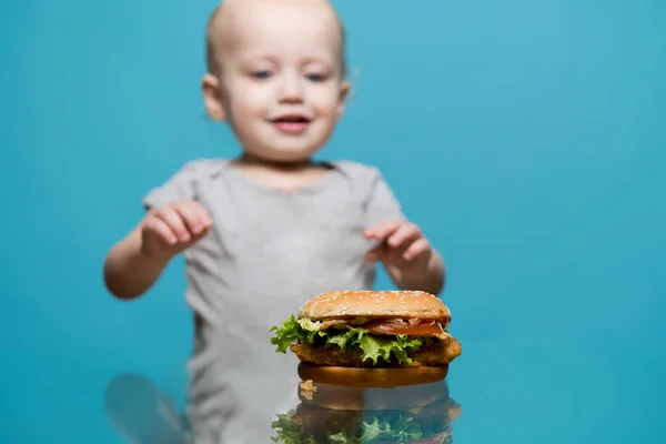 Małe dziecko zbliża hamburgera, który leży na szklanym stole — Zdjęcie stockowe