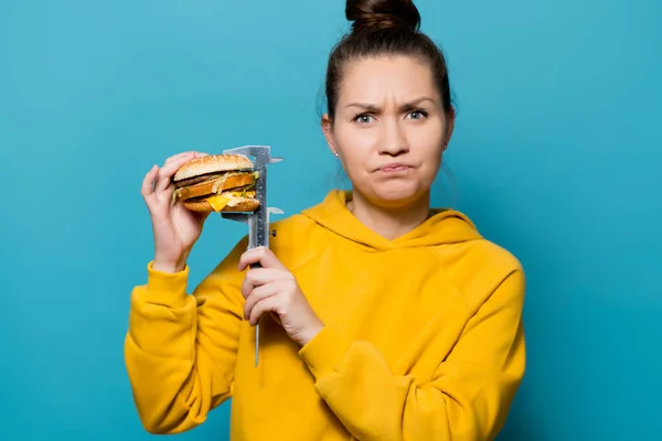 Het meisje toont ontevredenheid, het meten van de hoogte van de hamburger met een schuifmaat aan de zijkant van zichzelf — Stockfoto