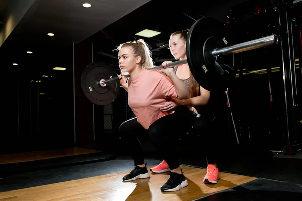The girl performs squats with a barbell, and the coach insures her — Stockfoto