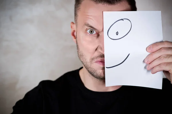 Wütender Mann bedeckt sein halbes Gesicht mit einem Blatt Papier mit einem gemalten Lächeln — Stockfoto