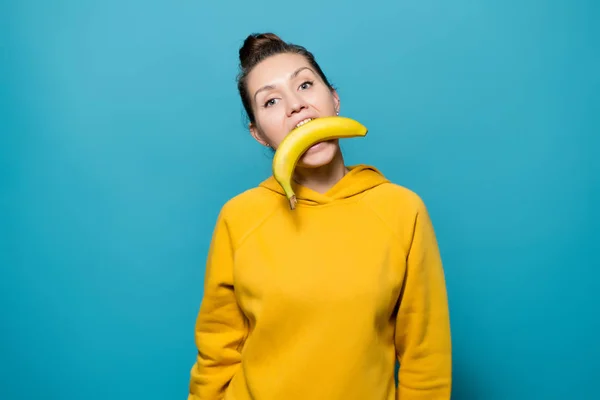 Een meisje in een helder sweatshirt houdt een banaan met haar tanden — Stockfoto