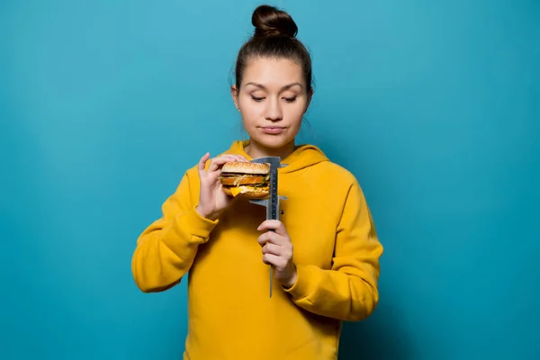 Een meisje in een sweatshirt meet de hoogte van een hamburger met een caliper — Stockfoto