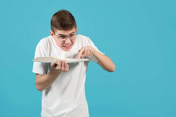 En nörd med glasögon tittar på kameran, håller i en bok och pekar finger åt den. — Stockfoto