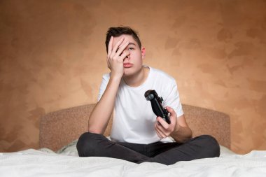 Delikanlı üzgün, yüzüne avuç içi yapıyor, ikinci elinde de bir joystick var.