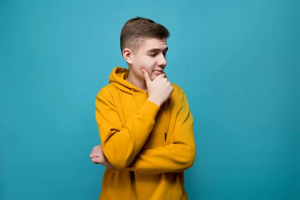 The young man in a yellow sweatshirt on a blue background is thoughtfully looking away — 스톡 사진