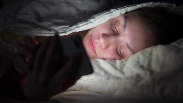 Chica ríe leyendo mensajes en el teléfono por la noche, acostado debajo de las sábanas — Vídeos de Stock