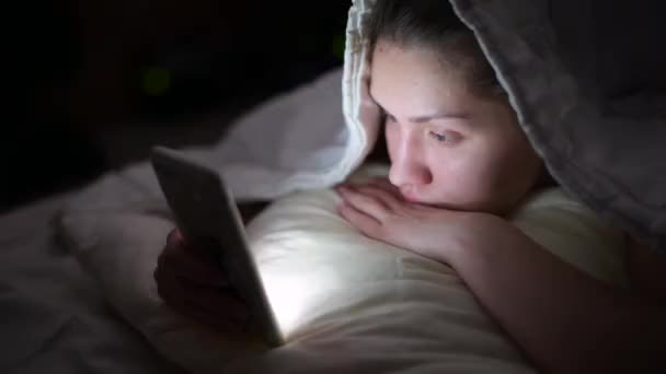 A girl without makeup at night looks at something on the phone and smiles. — Stock Video