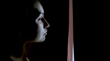 Karanlık bir odada üzgün bir kız karanlık bir perde açar ve pencereden dışarı bakar.