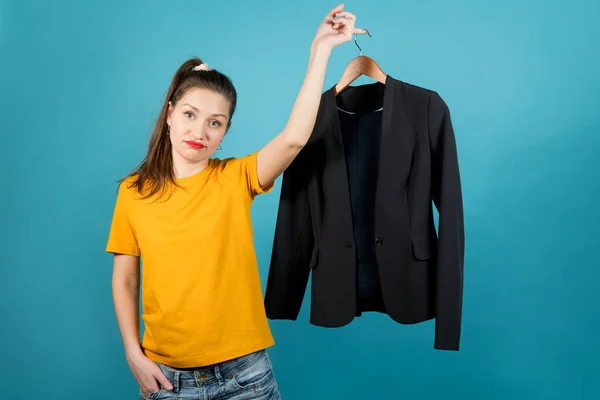 A young woman in bright clothes with sadness holds in her hand a hanger with strict office clothes