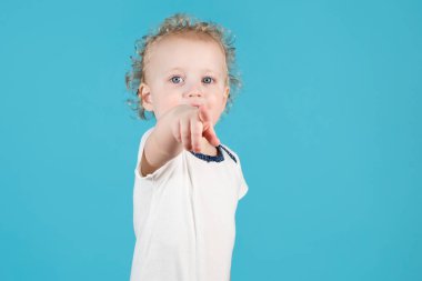 İki yaşındaki kıvırcık kız çocuğu izleyiciye parmağını sokuyor. Kamerada çocuk görünüyor. Mavi üzerine izole edilmiş.