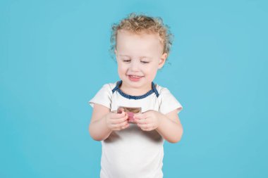 İki yaşındaki kıvırcık kız çocuğu elinde bir prezervatif tutuyor ve ona bakıp gülümsüyor. Mavi üzerine izole edilmiş.