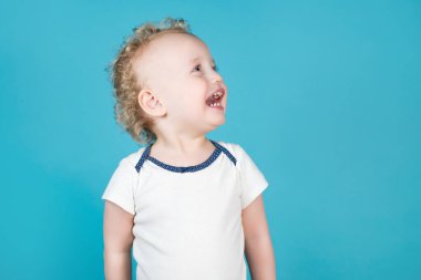 İki yaşındaki kıvırcık kız yan tarafa bakıp gülüyor. Mavi üzerine izole edilmiş.
