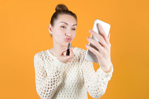 girl is going to paint her lips looking in the phone. Isolated on orange-yellow