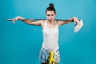 Temizlikçi ellerini paspasın üzerine asarak paspası omuzlarında tutuyor ve kameraya hoşnutsuz ve üzgün bakıyor. Kemerinde bir sprey ve bir bez asılı..