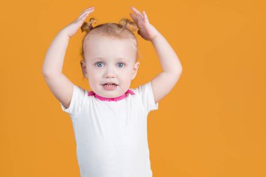 Küçük bir kız, şirin at kuyruğuyla elleriyle kafasına dokunuyor. Turuncu sarı arka planda izole edilmiş..