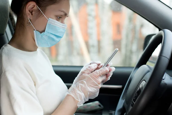 Woman in a medical mask and medical gloves holds a phone sitting in a car. Close up, side view