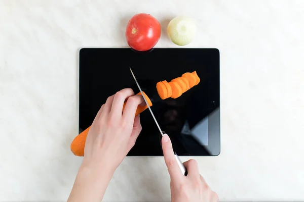 A woman cuts vegetables with a knife on an old graphics tablet, which serves as a cutting board. Top view