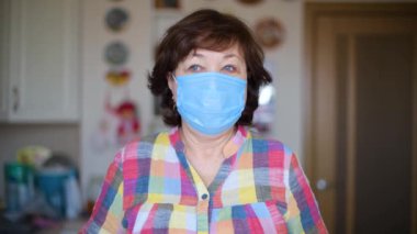 Koruyucu tıbbi maskeli yaşlı bir kadın çerçeveye bakıyor..