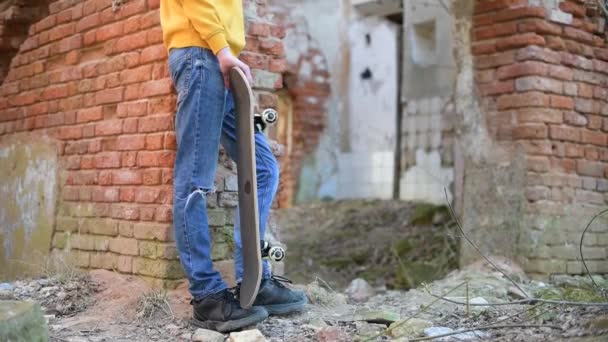 Um adolescente com um skate fica perto da parede de um edifício em ruínas — Vídeo de Stock