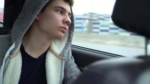 A young guy in a hood looks out the window, sitting in the back seat — Stock Video