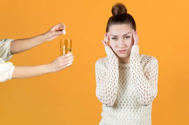 Genç bir kadın kafasına yapışır ve acıdan acı çeker. Kız arkadaşı onu coşkun bir tableti eritmeye ve acıyı dindirmeye davet ediyor. Turuncu sarı arka planda izole edilmiş.