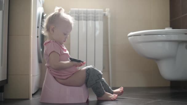 Una niña pequeña se sienta en una olla en el baño y sostiene un teléfono en sus manos — Vídeo de stock