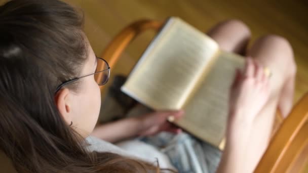 Una ragazza con gli occhiali sta leggendo un libro alla luce del giorno. Focus selettivo sugli occhiali . — Video Stock