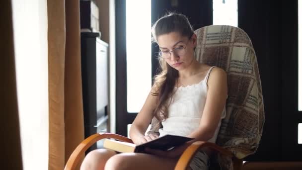 Giovane ragazza in occhiali legge un libro in luce naturale su una sedia a dondolo — Video Stock