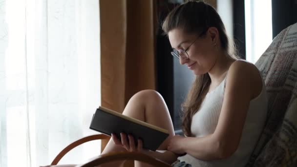 Gadis berkacamata membaca sebuah buku dengan cahaya alami di dekat jendela dan terkekeh. — Stok Video