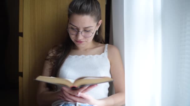 Ragazza con gli occhiali legge un libro vicino alla finestra e beve tè o caffè — Video Stock